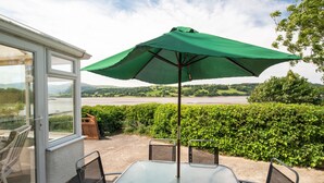 A patio with a table and chairs under a green umbrella, overlooking a river and hills.
