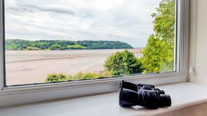 A window with a pair of binoculars on the ledge, overlooking a river and green hills.