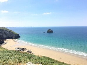 A coastal landscape with a sandy beach, a grassy cliff, and a rock formation in the sea. - 1 Jubilee Cottage (Tintagel)