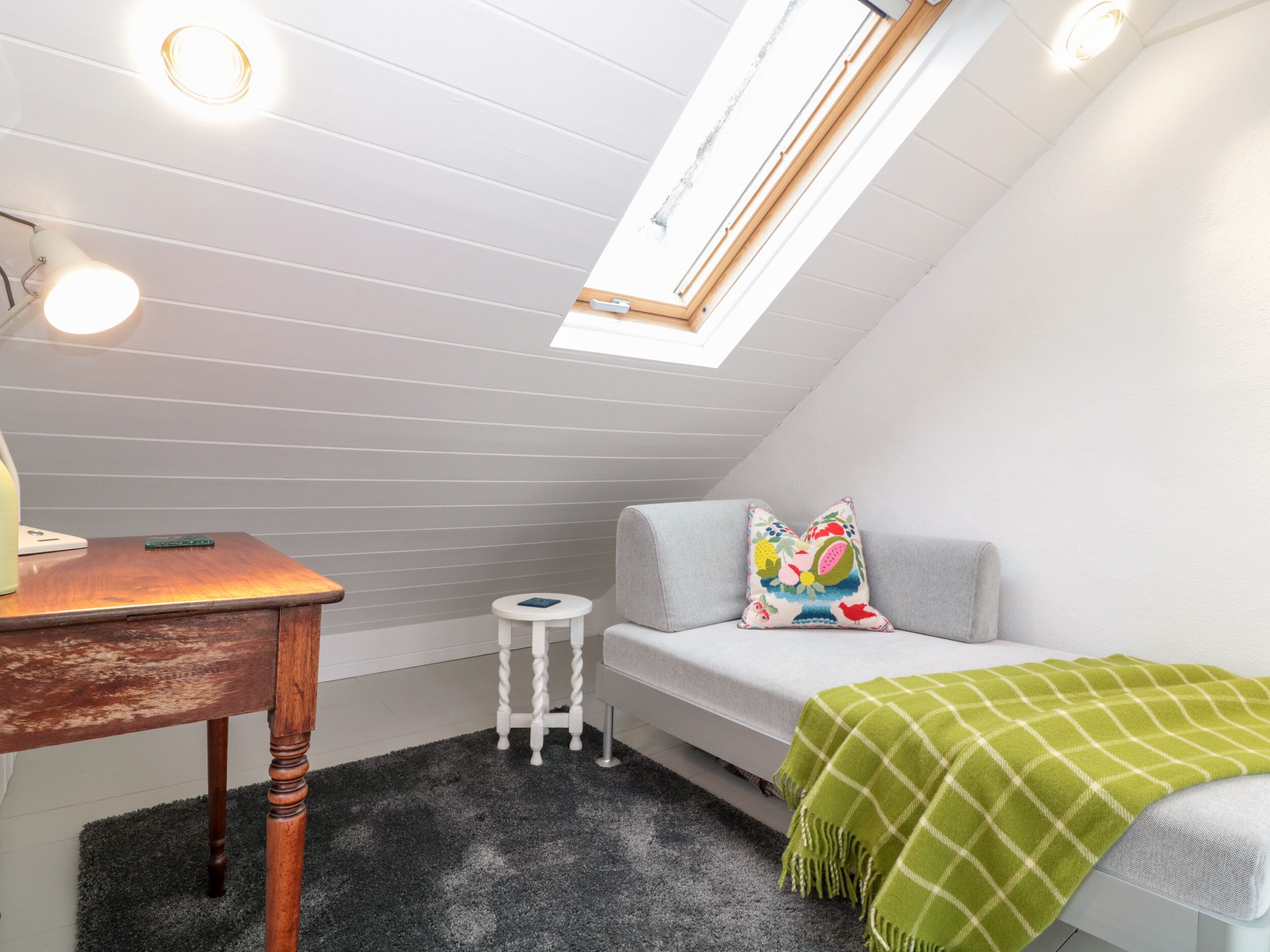 A small, cozy room with a sloped ceiling, a wooden desk, a white chair, a small round table, a grey sofa with colorful pillows, and a green checkered blanket.