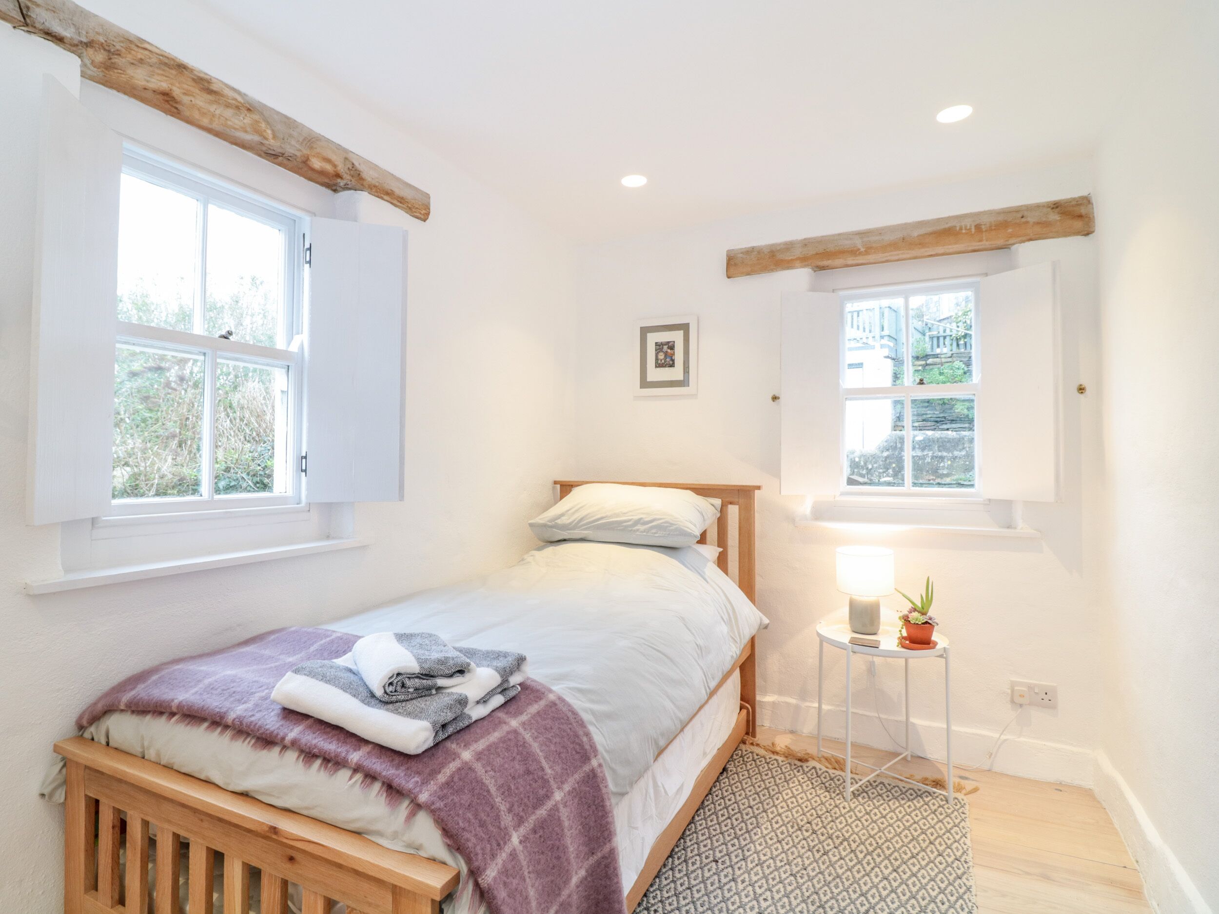 A bedroom with a bed, a small bedside table with a lamp, and two windows.