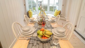 Un coin repas avec une table dressée pour six personnes, entouré de chaises blanches, une baie vitrée offrant une vue et un mur blanc orné d’un tableau.