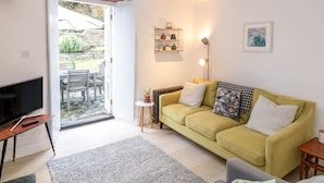 Un salon avec un canapé jaune, un téléviseur et une petite table. Une porte donne sur un espace extérieur aménagé avec une table et des chaises.