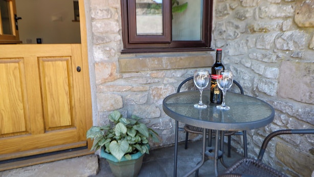 Un mur de pierre, une porte en bois, une table en verre avec deux verres à vin et une bouteille, et une plante en pot.