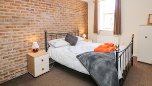 Une chambre avec un mur en briques, un lit recouvert d’une couverture grise, une table de chevet avec une lampe et une fenêtre avec des rideaux.