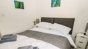 A neatly made bed with white and grey linens, a grey headboard, and a grey patterned throw. There are two small bedside tables with lamps, a white nightstand, and a wall-mounted power outlet. - Teàrlach House (Alnwick)
