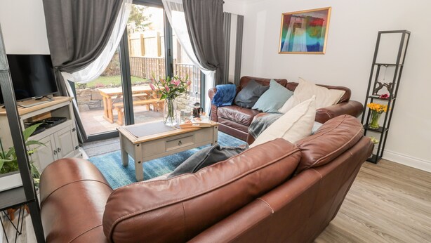 A living room with a brown leather sofa, a coffee table, a TV, and a dining area with a wooden table and chairs. - Teàrlach House (Alnwick)