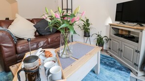 A living room with a brown leather sofa, a coffee table with breakfast items, a TV on a wooden stand, and potted plants. - Teàrlach House (Alnwick)