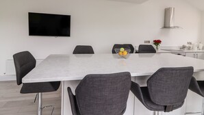 A modern dining area with a large table, six chairs, a flat-screen TV mounted on the wall, and a kitchen area with a stainless steel range hood. - Coolnaharragill Lower (Glenbeigh)