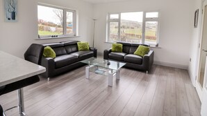 A modern living room with a black leather sofa, two green cushions, a glass coffee table, and large windows offering a view of the countryside. - Coolnaharragill Lower (Glenbeigh)