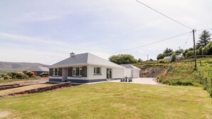 A single-story house with a sloped roof, a front porch, and a stone retaining wall. - Coolnaharragill Lower (Glenbeigh)