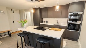 A modern kitchen with a large island, two bar stools, a built-in oven, and pendant lighting. - The Old Workshop (Denbigh)