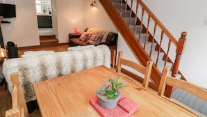 A cozy living room with a wooden dining table, a fluffy white sofa, a wooden staircase, and a television mounted on the wall.