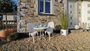A stone house with a small patio area featuring a table and chairs, a potted plant, and a decorative sphere. - Valley View (St Austell)