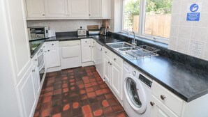 A compact kitchen with white cabinets, a black countertop, a built-in washing machine, and a window with a view of a wooden fence. - Ty'r Winllan (Newport)