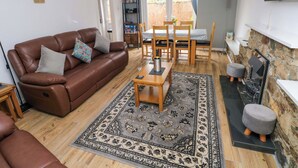 A cozy living room with a brown leather sofa, a wooden coffee table, and a patterned rug. There is a dining area with wooden chairs and a table, and a stone fireplace. - Ty'r Winllan (Newport)