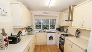 A compact kitchen with a washing machine, microwave, toaster, and wooden flooring. - 4 Ty Rhosyn (Porthmadog)