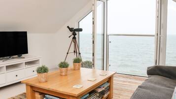 Un salón moderno con un ventanal grande con vista al mar, una mesa de centro de madera con libros, un televisor y una cámara sobre un trípode.
