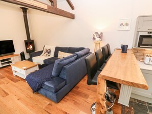 A modern living room with a dark blue sectional sofa, a wooden coffee table, a TV, a fireplace, and a kitchen area with a microwave.