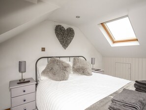 Ein Schlafzimmer mit schrägen Wänden, einem Dachfenster, einem Bett mit flauschigen Kissen, einem Nachttisch mit Lampe und einer Wanddekoration in Herzform.