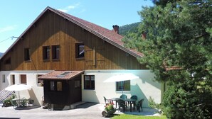 A two-story wooden building with a tiled roof, a covered patio area with outdoor seating, and a large tree in the foreground. - Cozy Mountain Retreat with Garage, BBQ & Wifi in La Bresse (La Bresse)