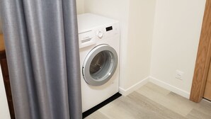 A compact, white Bosch washing machine in a small laundry room with light wooden flooring and a grey curtain partially drawn. - Cozy Mountain Retreat with Garage, BBQ & Wifi in La Bresse (La Bresse)