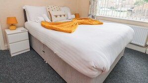 Uma cama bem arrumada com edredom branco e manta amarela, uma luminária de cabeceira, um criado-mudo pequeno e uma janela com cortinas.