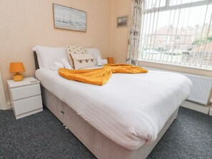 A neatly made bed with a white comforter and a yellow throw blanket, a bedside lamp, a small nightstand, and a window with curtains.