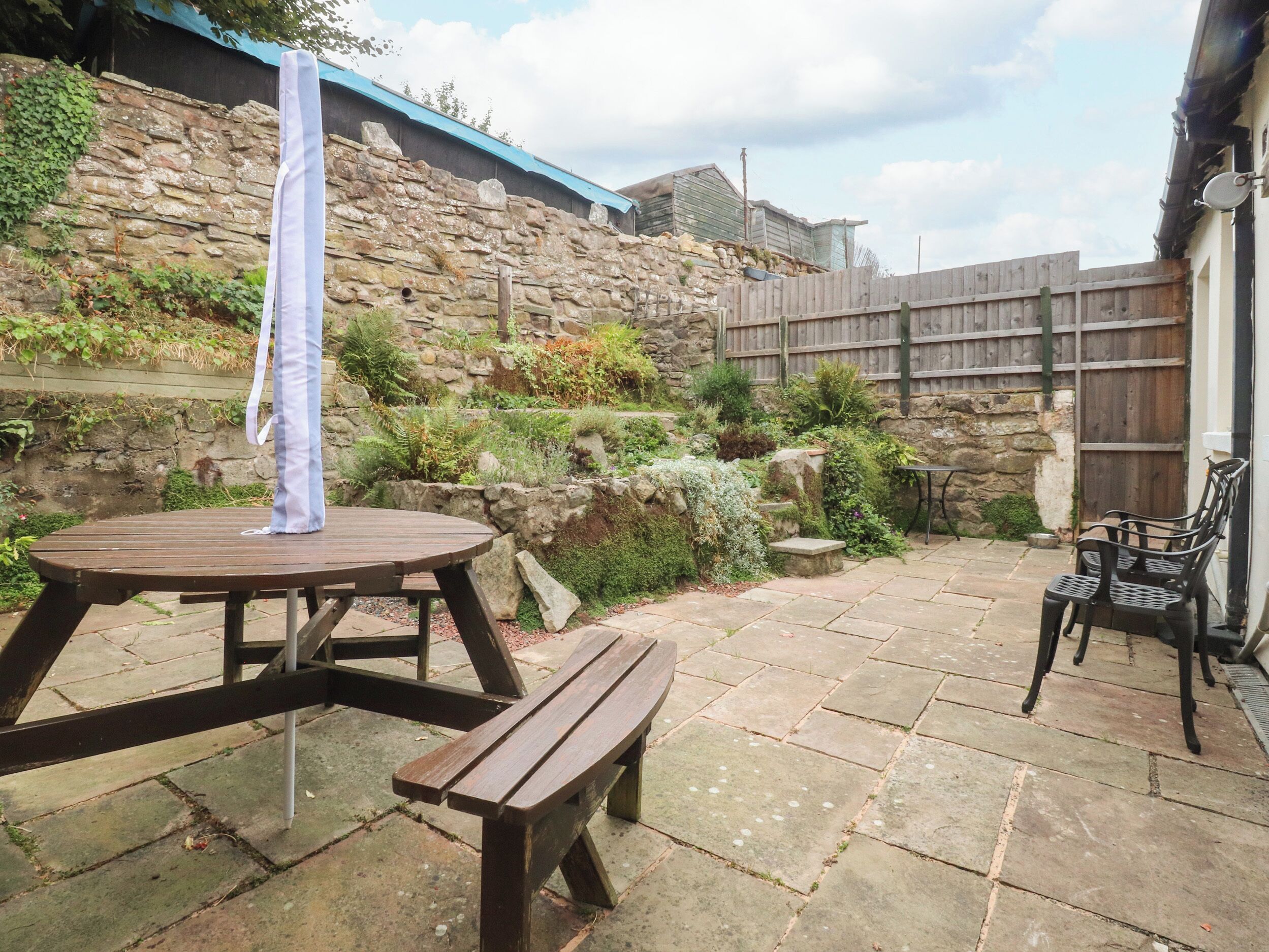 A patio with a wooden table and bench, surrounded by stone walls and greenery.
