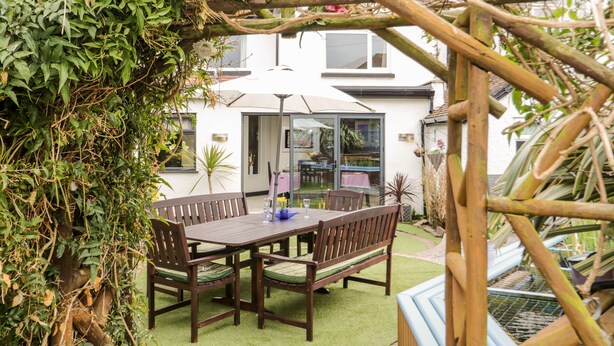 Eine Terrasse mit Holztisch und -stühlen, umgeben von Grünpflanzen und einer Pergola.