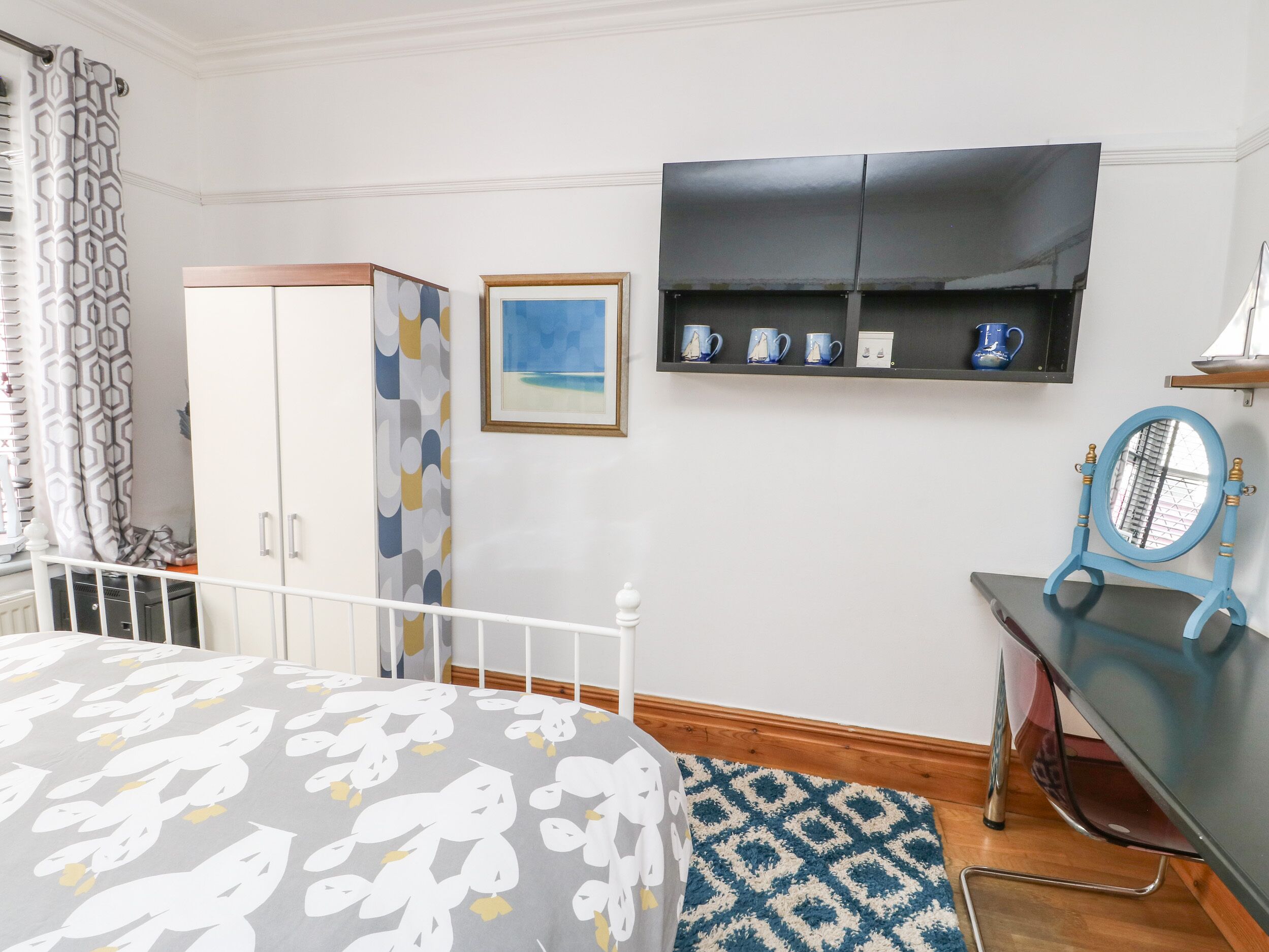 A bedroom with a bed, a wardrobe, a desk with a mirror, and a shelf with decorative items.