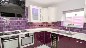 Eine moderne Küche mit violetten und weißen Schränken, Edelstahlgeräten und einem Fenster mit rosa Lamellen.