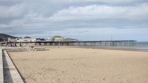 Ein Strand mit einem Steg, der sich ins Wasser erstreckt, einem Sandbereich und einem Gebäude im Hintergrund.