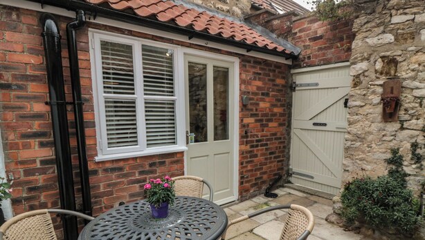A brick house with a white door and shutters, a small patio with a round table and wicker chairs, and a stone wall. - Dove Cottage (Pickering)