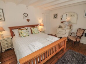 Un dormitorio con cama de madera, ropa de cama blanca, una mesita de noche con lámpara, cómoda, silla y espejo.