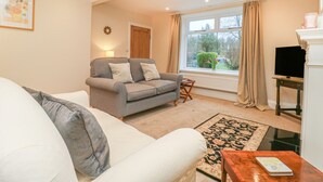A living room with a grey sofa, a white sofa, a small table with a book, a TV on a stand, and a window with curtains. - Westholme (Barnard Castle)