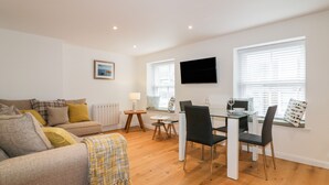 A modern living room with a sofa, a dining table with chairs, a TV mounted on the wall, and a window with blinds. - Midships (St Austell)