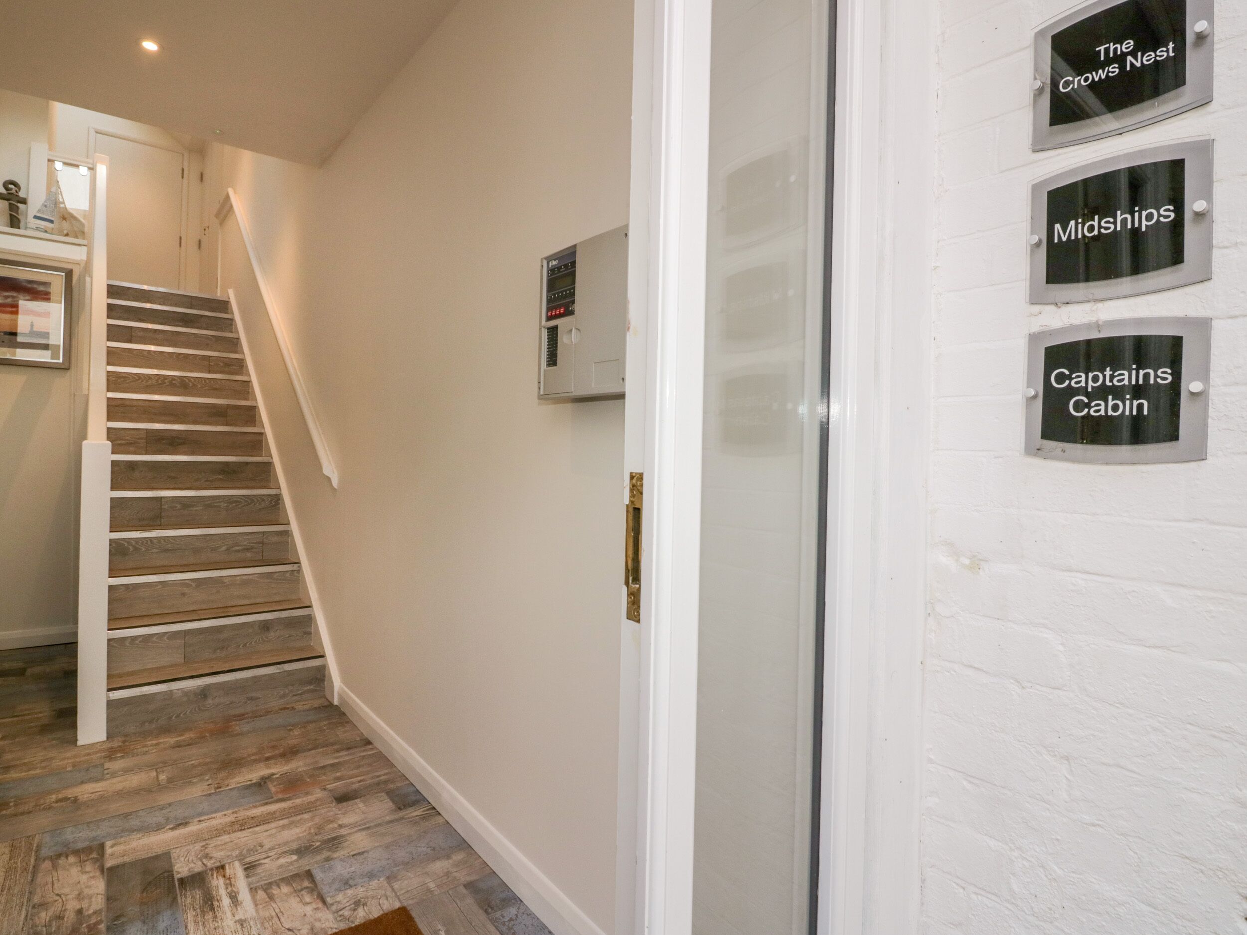 A staircase leading to a landing with a wall-mounted thermostat and three signs indicating room names: 'The Crows Nest,' 'Midships,' and 'Captains Cabin.'