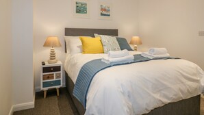 A neatly made bed with white and blue bedding, two bedside lamps, and a small bedside table with a lamp and a remote control. - Midships (St Austell)
