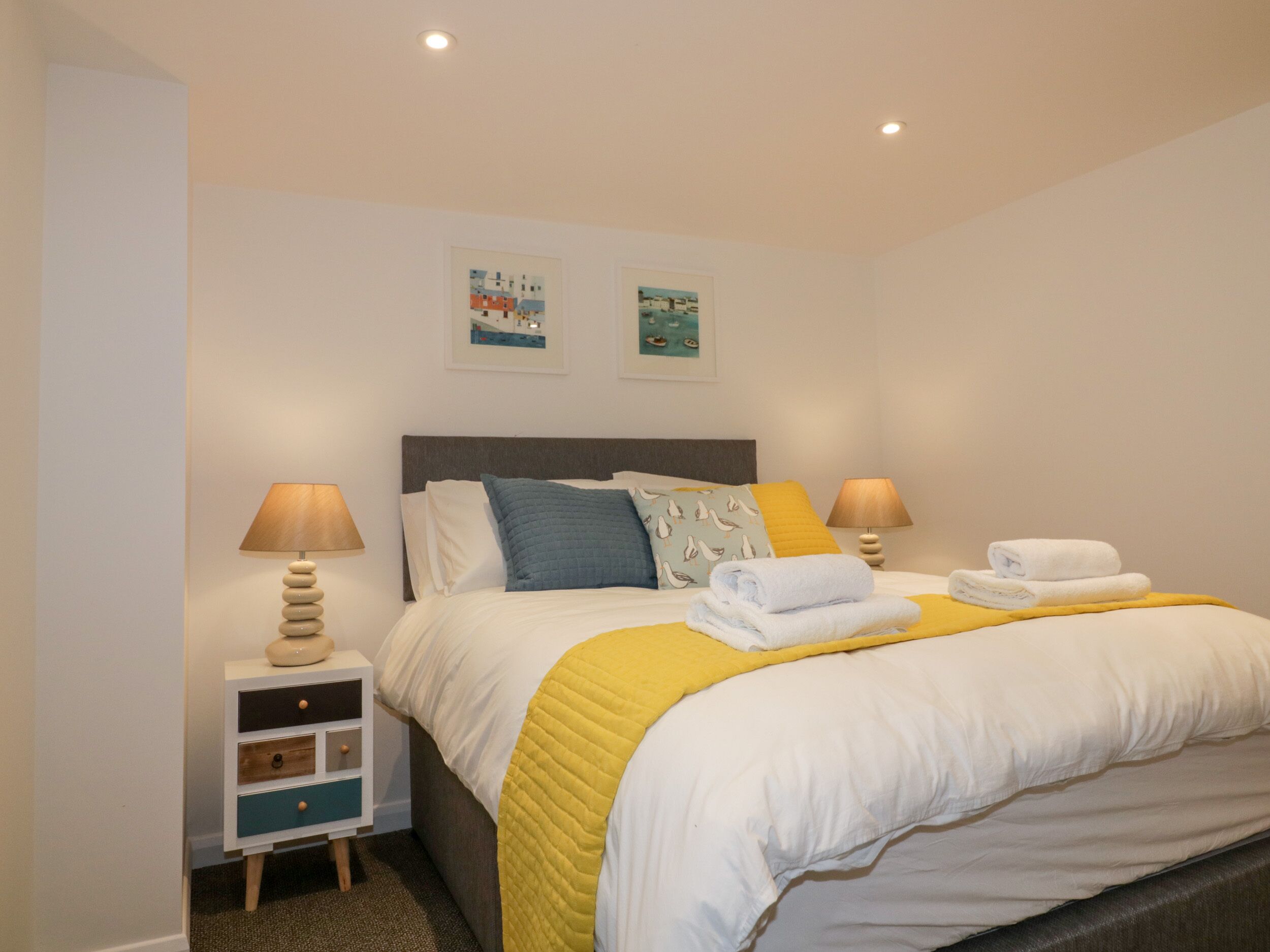 A neatly made bed with white and yellow bedding, two pillows, and folded towels on a bedside table.