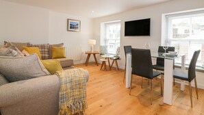 A modern living room with a sofa, a dining table with chairs, a TV mounted on the wall, and a window with blinds. - The Crows Nest (St Austell)