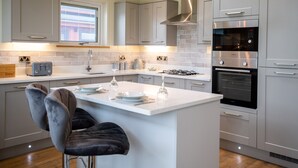 A modern kitchen with grey cabinets, a central island with bar stools, a built-in oven, and a microwave. - Otter Lodge (Skipton)