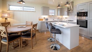 A modern kitchen with wooden flooring, a central island with bar stools, pendant lighting, and built-in appliances. - Otter Lodge (Skipton)