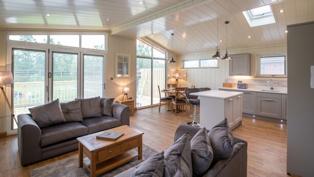 A spacious living area with a sofa, coffee table, and a dining table with chairs. There is a kitchen area with a sink and cabinets. - Otter Lodge (Skipton)