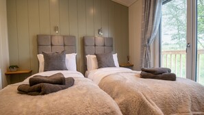 A double bed with fluffy blankets and pillows, a nightstand with a lamp, and a window with curtains. - Otter Lodge (Skipton)