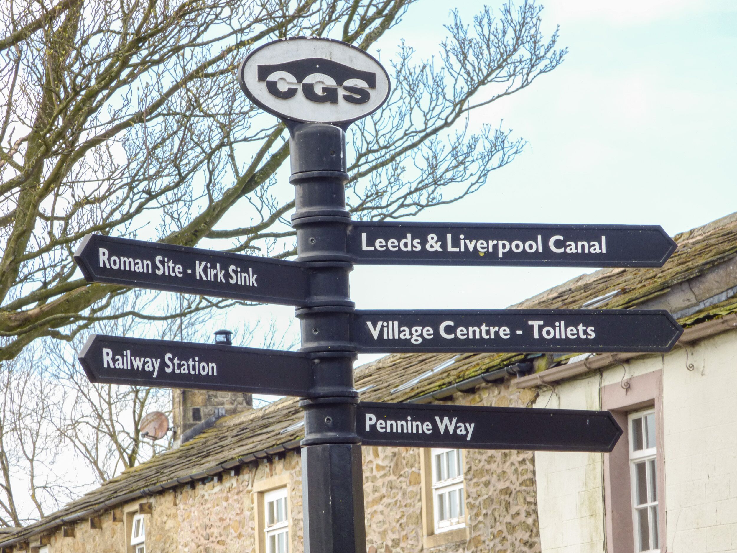 A directional signpost with multiple arrows pointing to various destinations including Roman Site, Kirk Sink, Leeds & Liverpool Canal, Village Centre, Toilets, Railway Station, and Pennine Way.