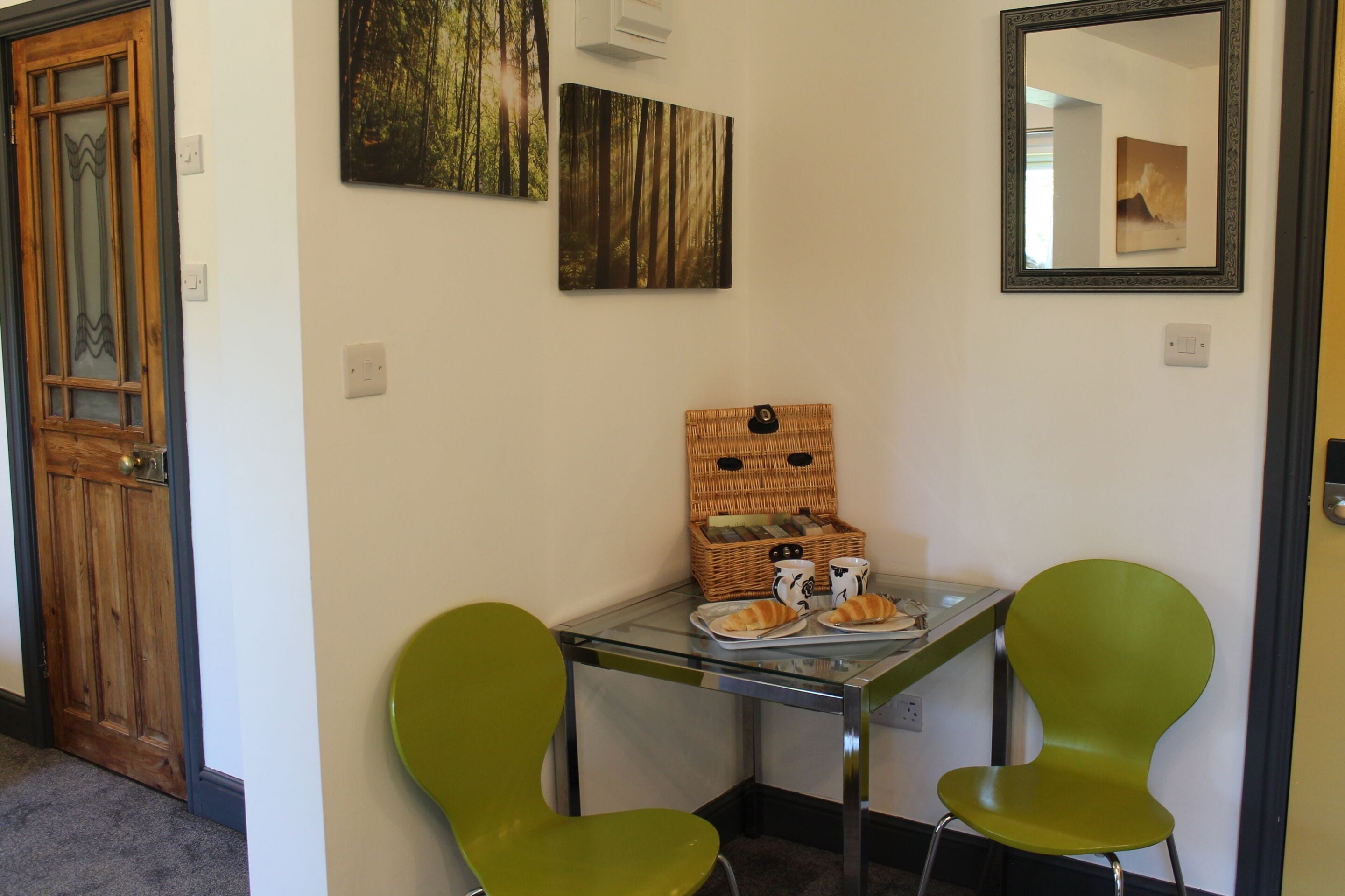 A room with a wooden door, a glass table with two green chairs, a wicker basket with books and a mug, and two framed pictures on the wall.