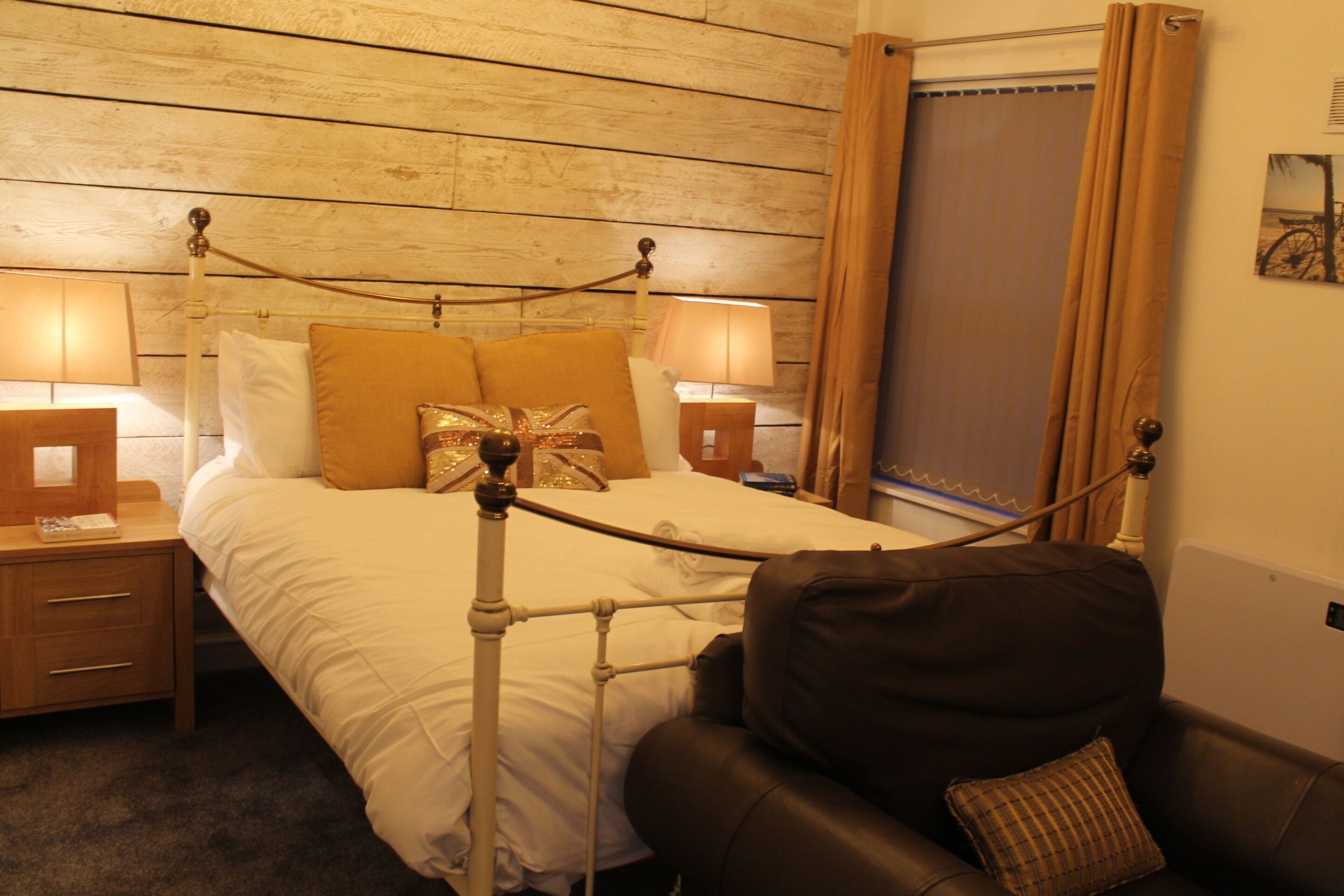 Une chambre à coucher avec une tête de lit en bois, un lit recouvert de draps blancs, deux lampes de chevet, un fauteuil en cuir marron et un rideau marron.