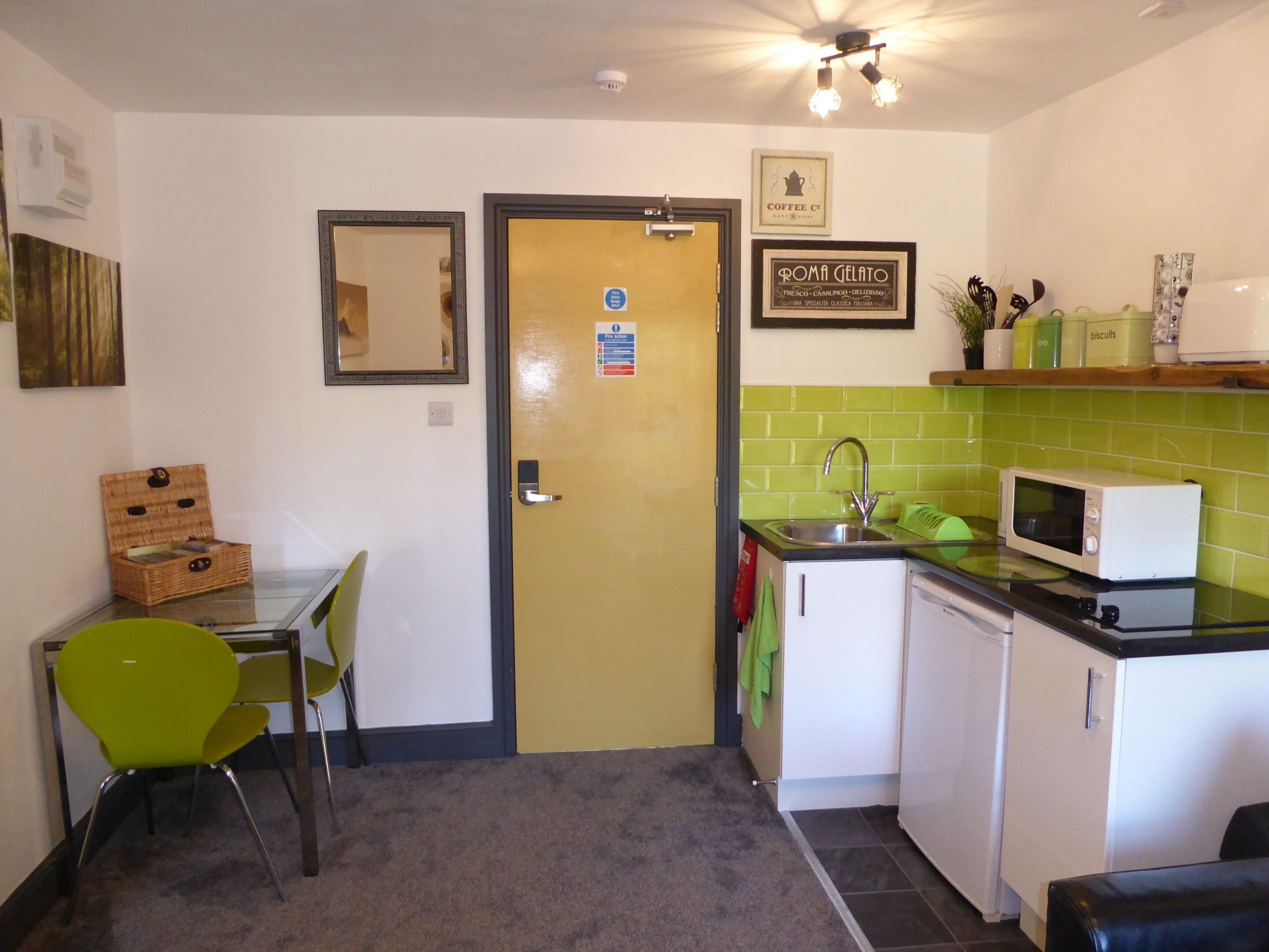 A compact kitchen with a microwave, sink, and a small refrigerator. There is a dining area with a chair and a table, and a door with a sign.