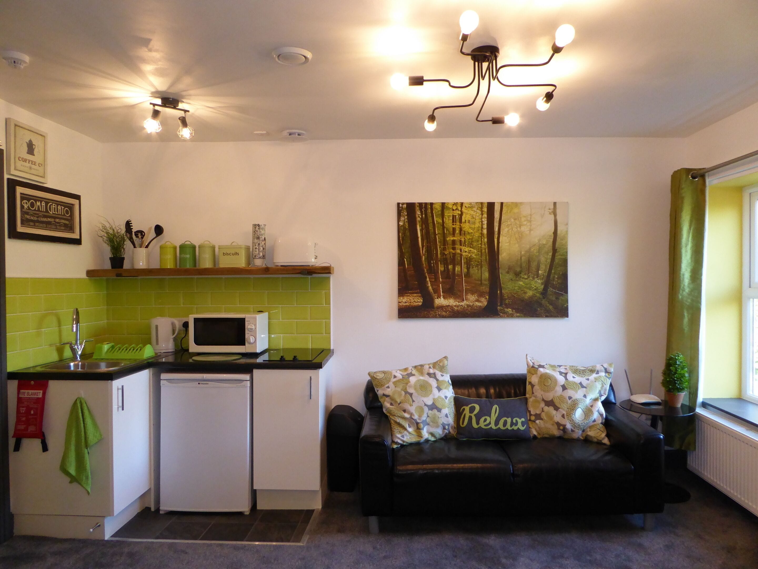 A compact kitchen with a microwave, a small refrigerator, and a sink. There's a green-tiled backsplash, a black leather sofa with 'Relax' pillows, and a forest-themed painting on the wall.